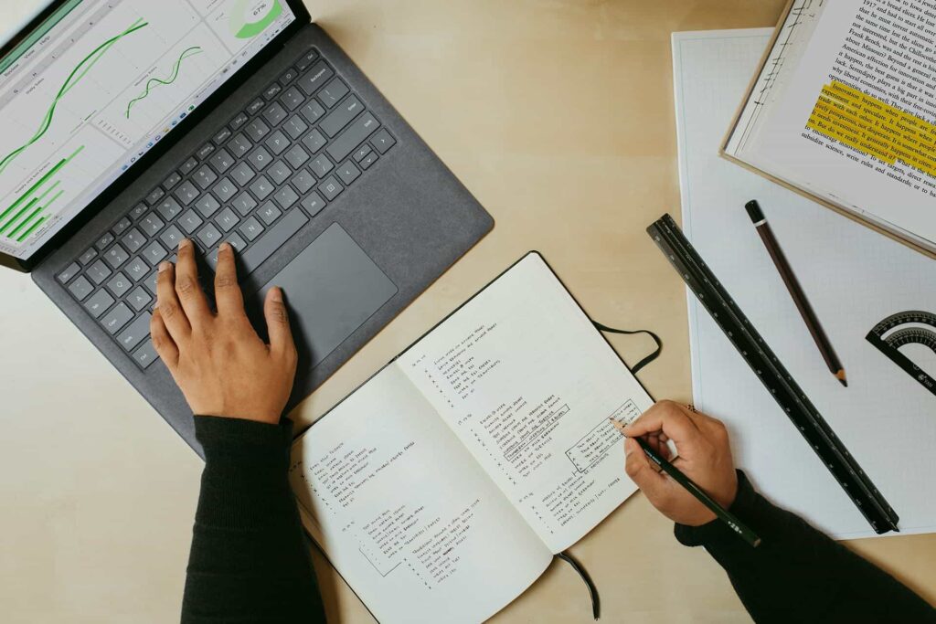Overhead view of hands using a laptop with charts and writing in an open notebook on a desk with a ruler, pencil, and highlighted book.