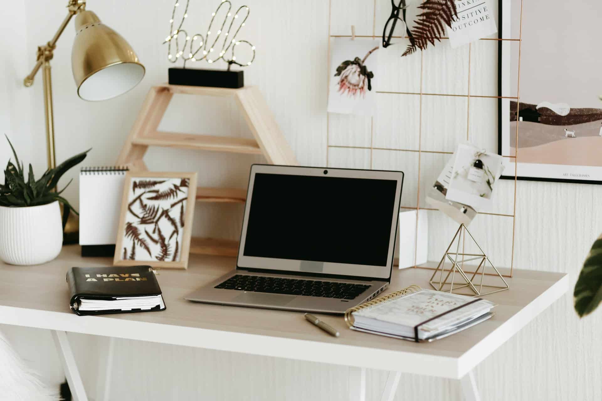 Bright desk setup with an open laptop, planner, notebook, desk lamp, plant, and decorative wall grid.