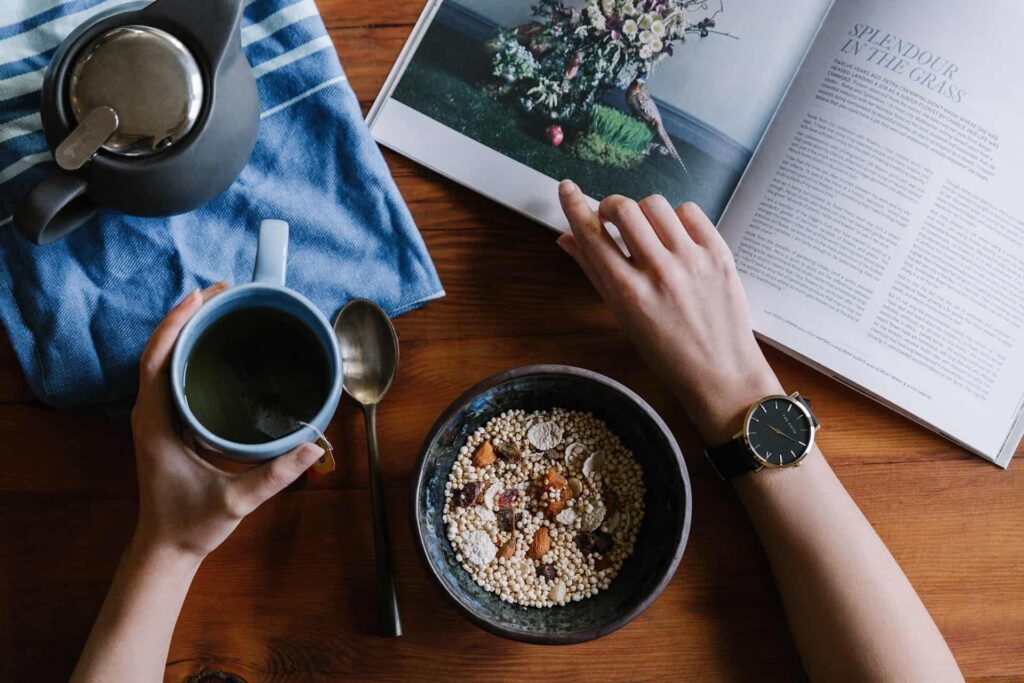Overhead view of a person holding a cup of tea beside a breakfast bowl and an open magazine on a wooden table.