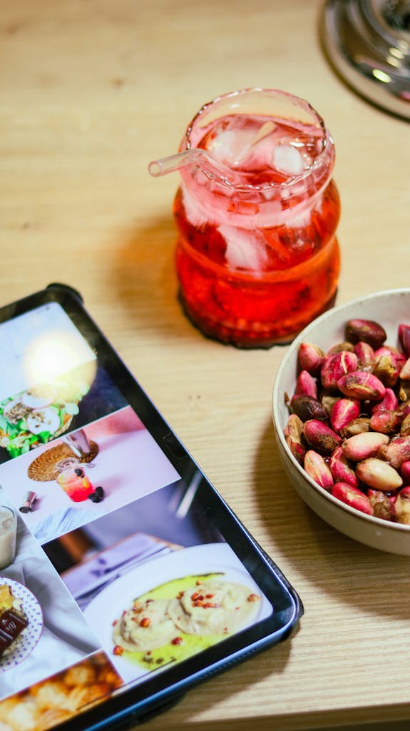 Tablet showing food and recipe images beside a red iced drink and a bowl of nuts on a wooden table.