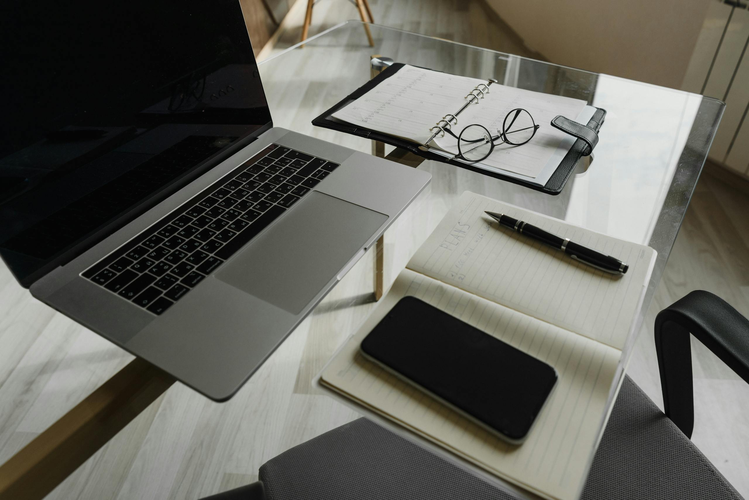 Clean and organized workspace with a laptop, notebook, and pen on a glass desk.
