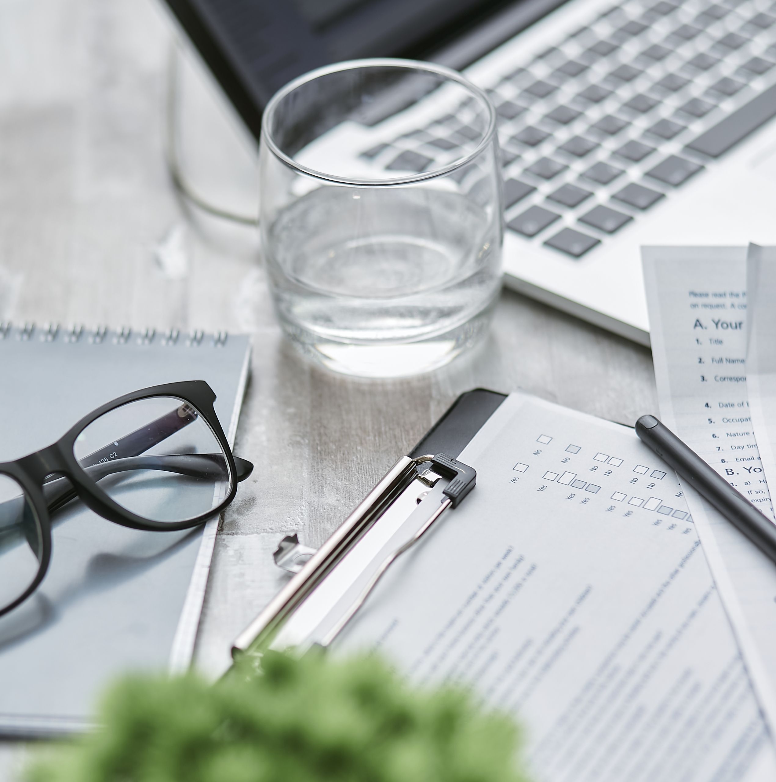 Close-up of a workspace with glasses, a laptop, papers, a clipboard, a pen, and a glass of water.