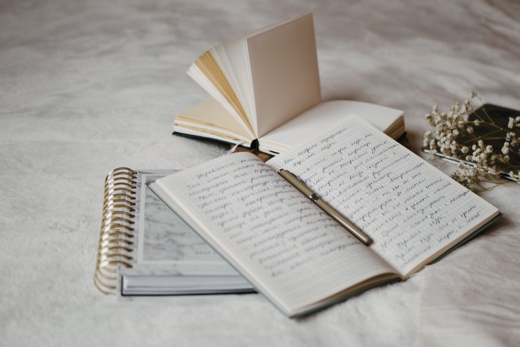Close-up of open notebooks with handwritten notes and a pen on a soft bed surface with delicate dried flowers.