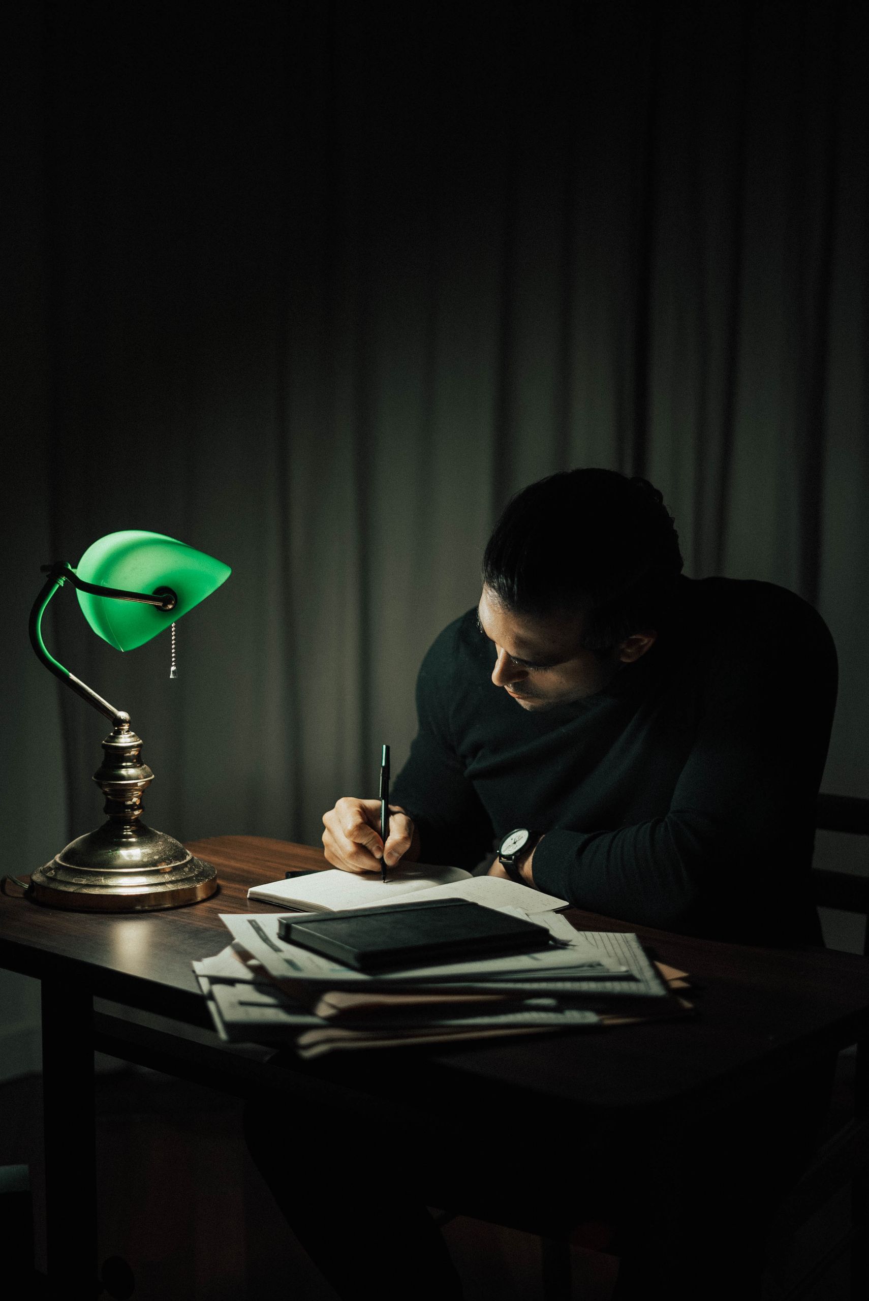 Focused man taking notes in a notepad at a desk with documents and a table lamp in a dark home office.