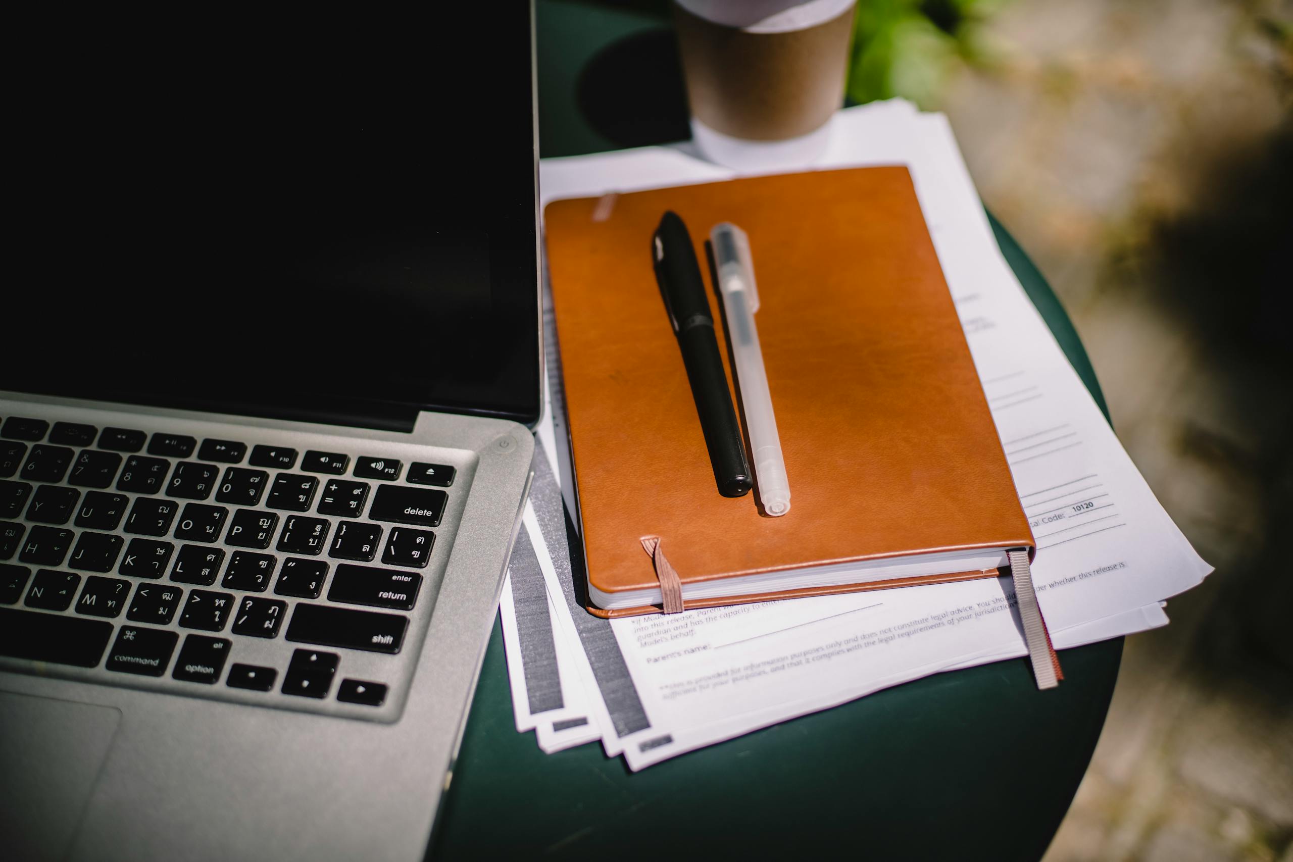 Overhead view of a leather notebook, papers, pens, a laptop, and a cup of coffee on a table.