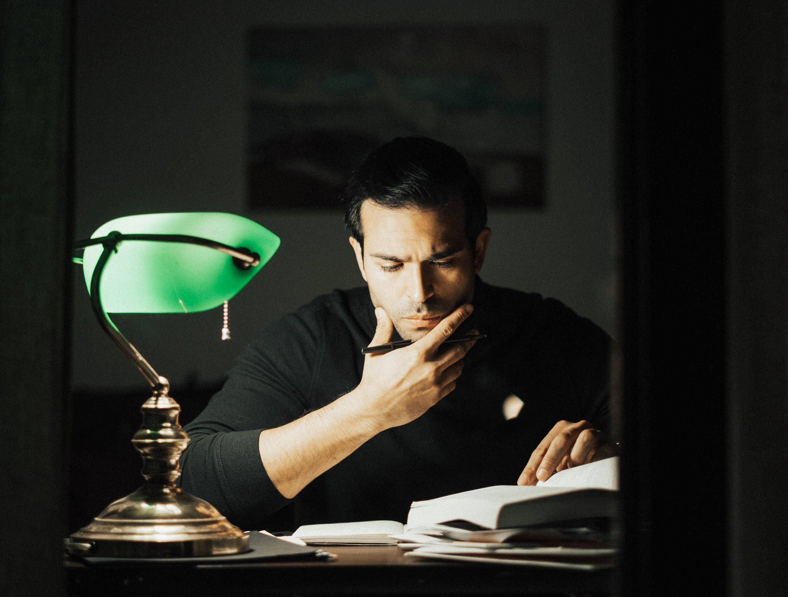 Young man in a black turtleneck reading a book and touching his chin while studying in a dark home office.